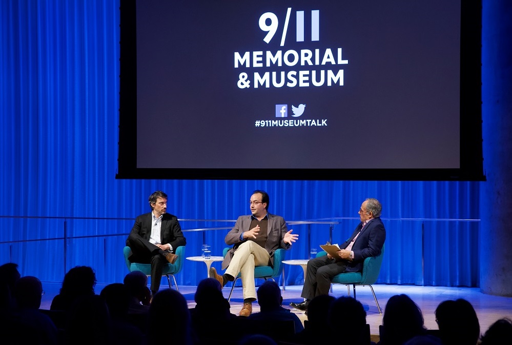 Three men in suits sit on a blue-lit auditorium stage. Behind them "9/11 Memorial & Museum #museumtalk" is projected on a screen behind them. The heads of the audience members appear in silhouette in the foreground.