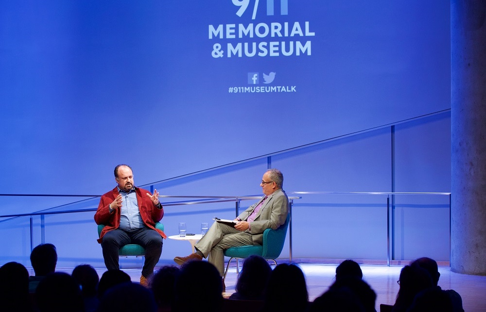 In this wide-angle photograph, two men sit on a blue-lit auditorium stage, with “9/11 Memorial & Museum #museumtalk” projected behind them. The heads of the audience members appear in silhouette in the foreground.