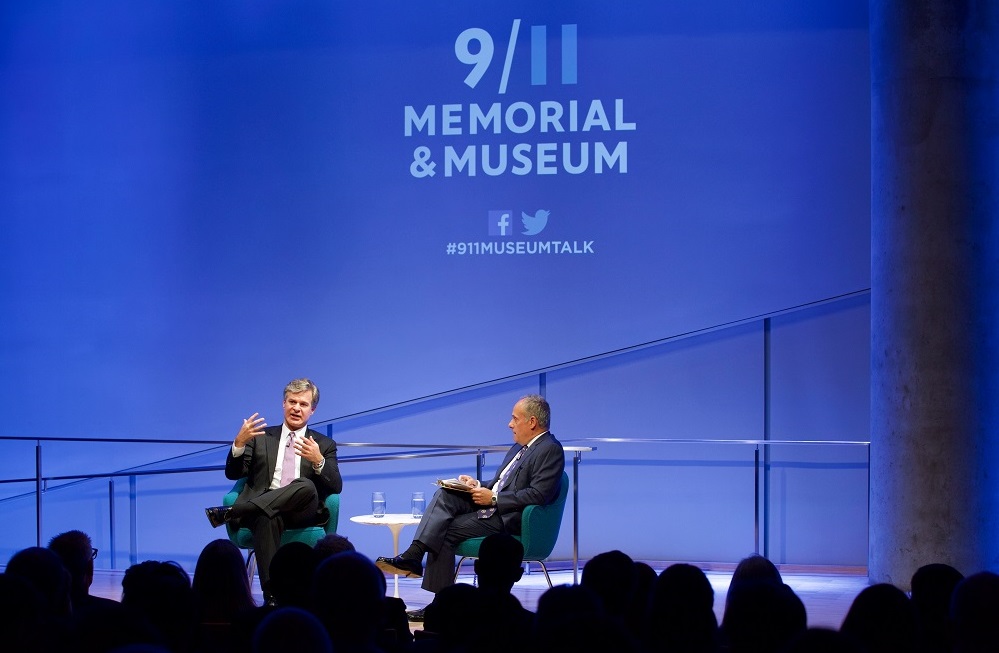 Two men in suits sit on a blue-lit auditorium stage with “9/11 Memorial & Museum #museumtalk” projected behind them. Heads of audience members appear in silhouette in the foreground.