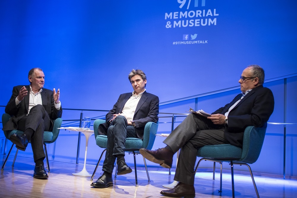 Three men, two speakers and a moderator, sit with crossed legs on a blue-lit auditorium stage during a 9/11 Memorial Museum public program.