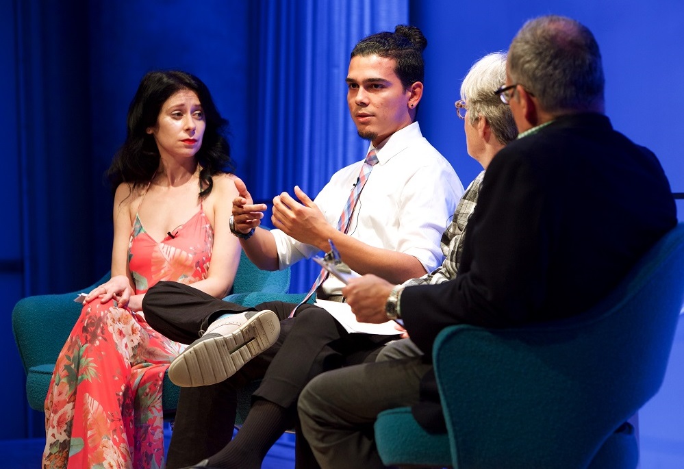 Four people sit on a blue-lit auditorium stage during a public program.