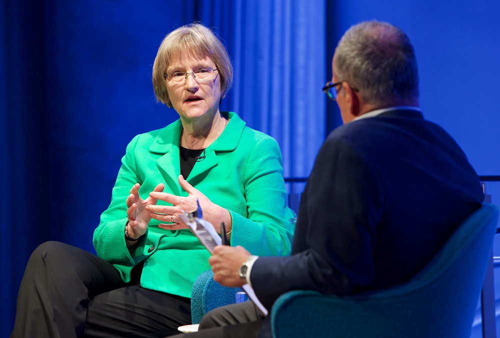 A woman in a green suit jacket speaks on a blue-lit auditorium stage to a moderator, whose back is to the camera.
