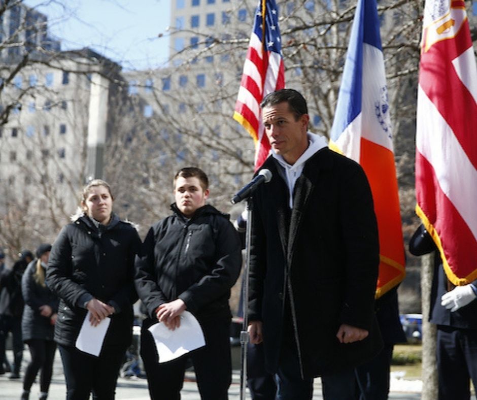 A man with dark hair in a dark coat stands at a microphone while a young woman and young man, also in dark coats, look on from the left