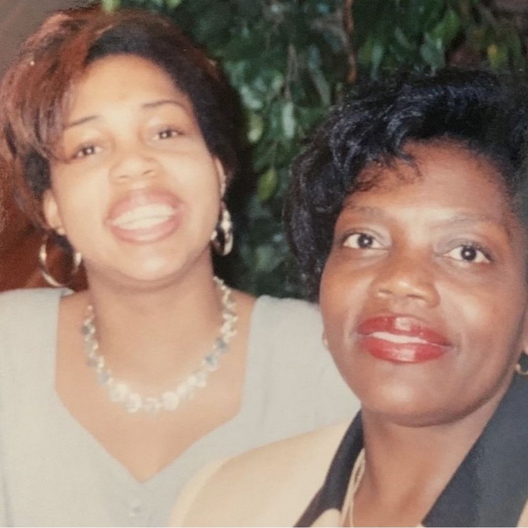Two dark-skinned women smile at the camera in front of a tree. The woman on the left is younger and wears a beige top; the woman on the right wears a black top and red lipstick.