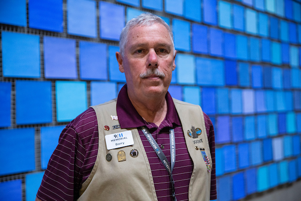A gray-haired man with a mustache, in a burgundy shirt and khaki volunteer vest, in front of blue tile-like installation.