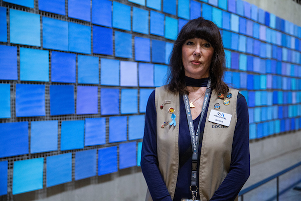 A brunette woman in a black shirt and khaki volunteer vest, in front of blue tile-like installation.