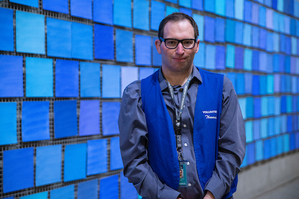 A young man with dark hair and glasses wearing a gray shirt and khaki volunteer vest, in front of blue tile-like installation.