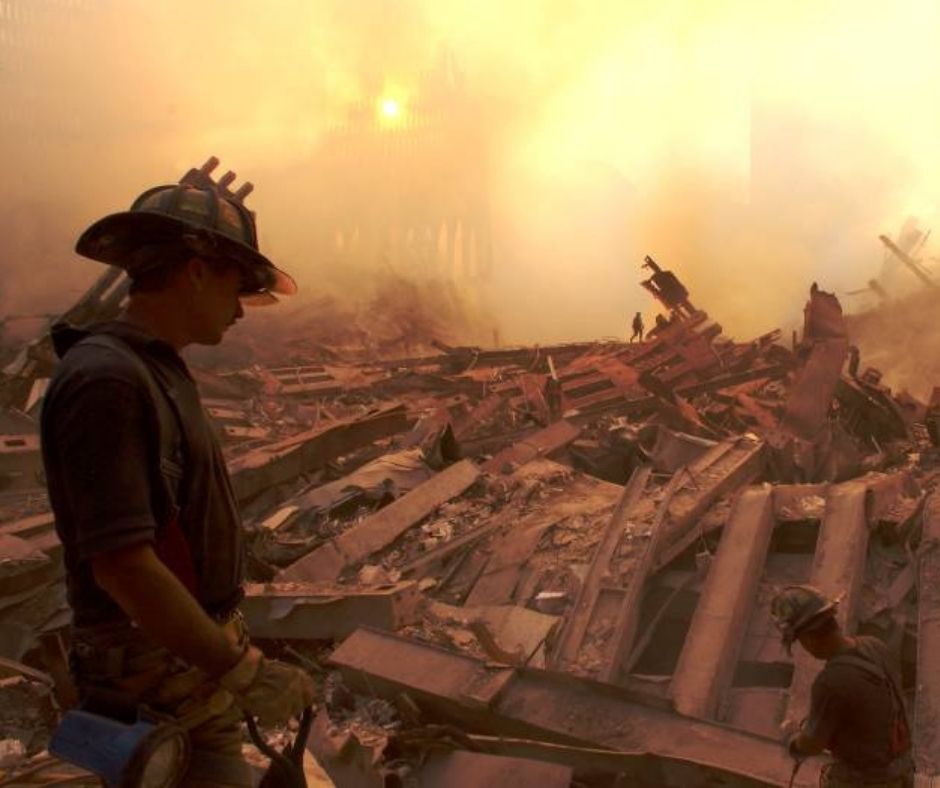 A rescue and recovery worker (lower left) looks out at the wreckage of Ground Zero, with an orange tinged sky 