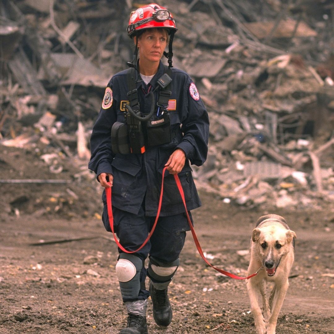 A female emergency worker in a dark blue uniform walks on the pile with a cream-colored shepherd dog on a leash