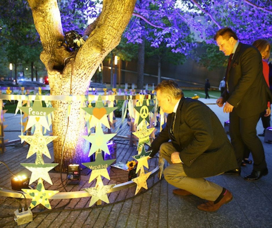 Diplomats, one in a dark jacket and khaki pants and one in a dark suit, crouch at the base of the Survivor Tree, where hand-painted wooden stars hang