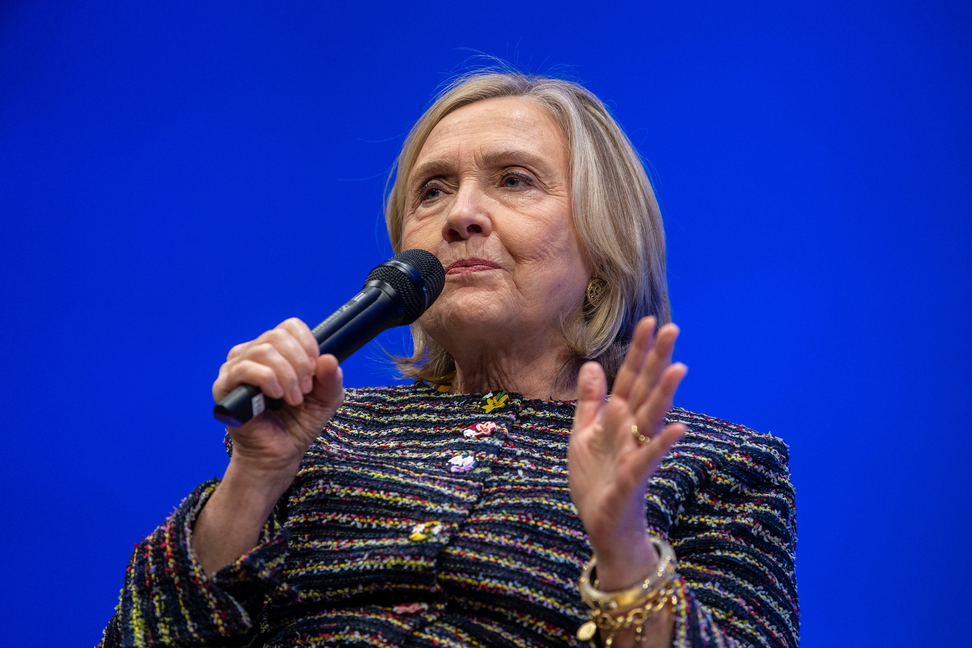 Hillary Rodham Clinton against a bright blue curtain, holding a microphone and speaking. She wears a patterned blazer.