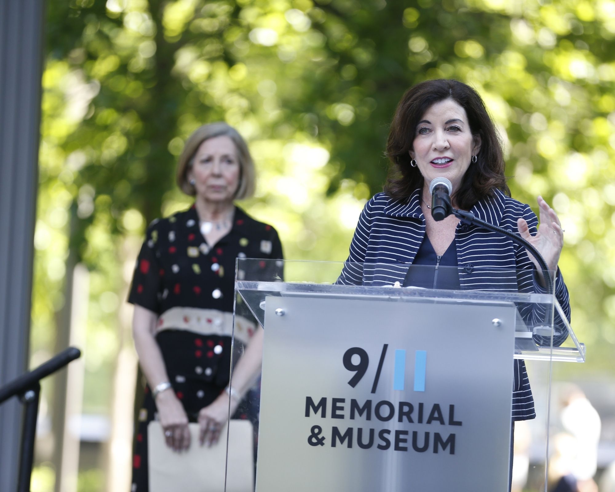 Alice Greenwald, our President & CEO, wears a dark dress and stand behind New York governor Kathy Hochul speaking at a podium