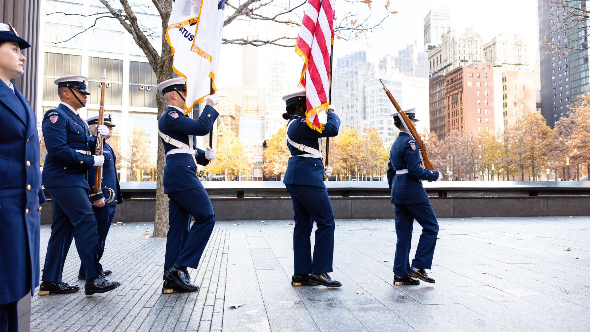 Four Coast Guard members in uniform walk across the plaza carrying instruments, flags, and a rifle, with the Memorial in background