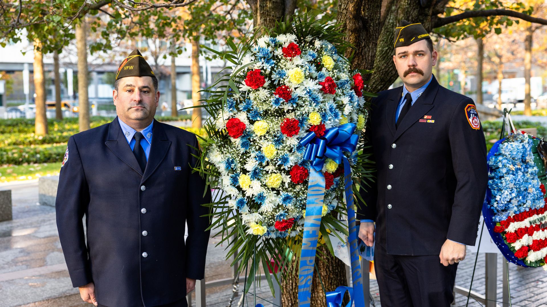 Two people in uniform stand at the Survivor Tree with a tribute wreath between them
