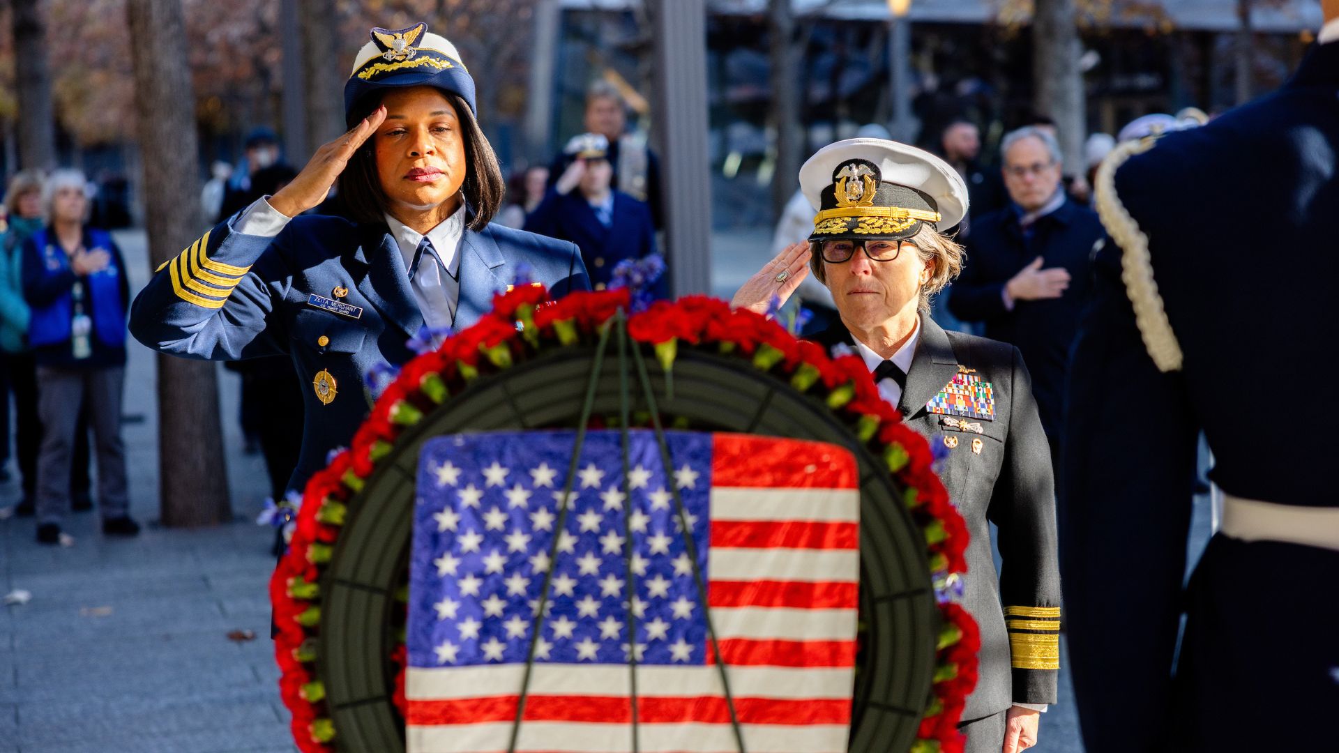 Two women in military uniforms salute a tribute wreath with an American flag at center