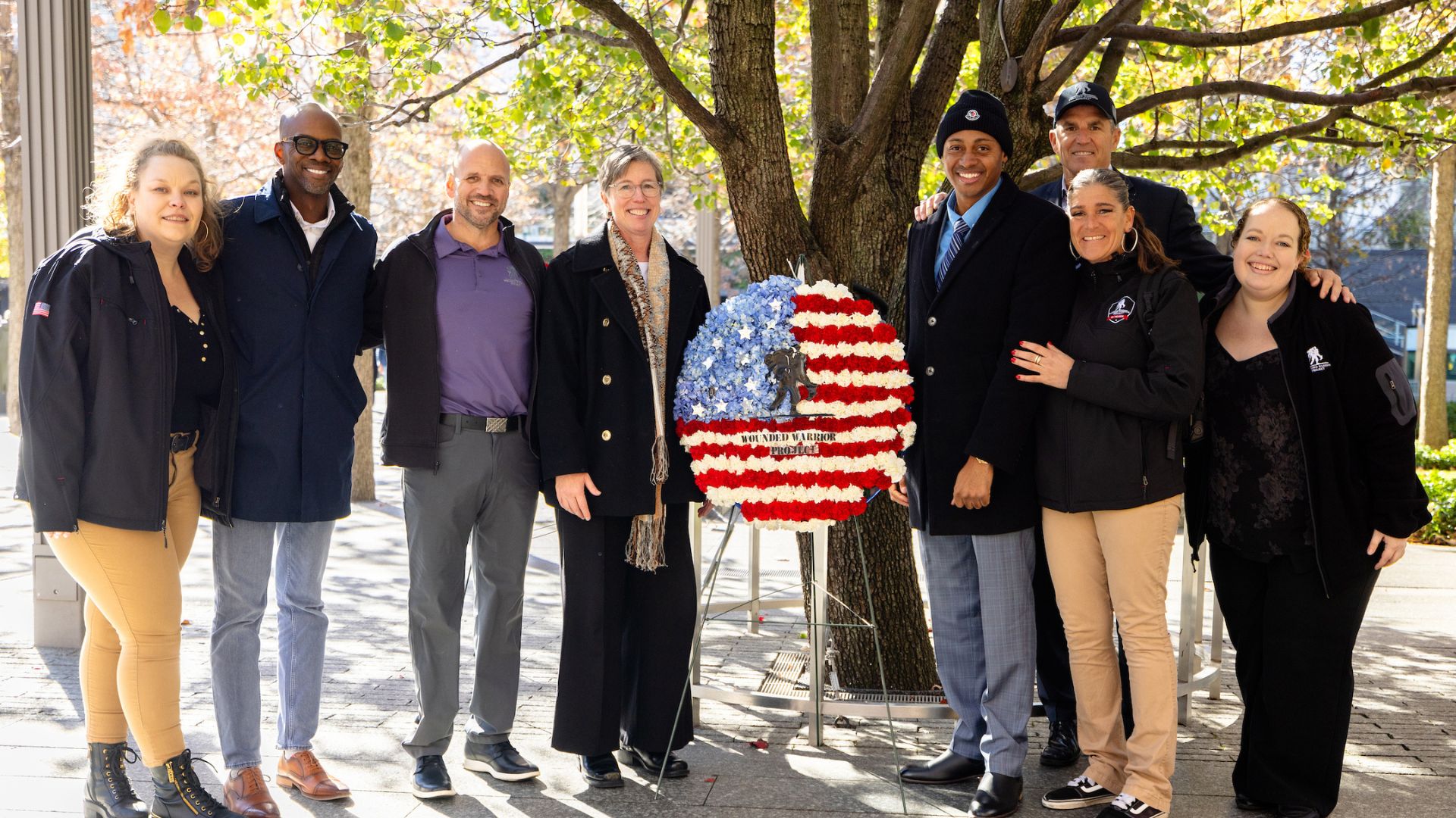 A row of smiling people at the Survivor Tree, with an American Flag wreath at center