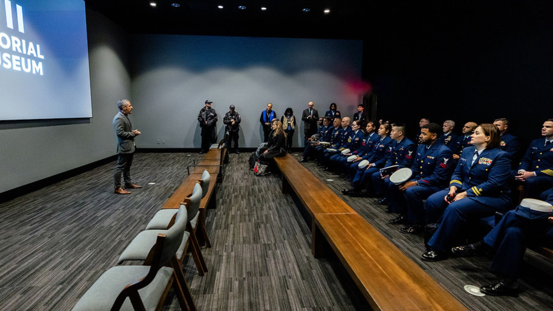  A group of uniformed Coast Guard members in the auditorium, listening to a staff member speak in front of a movie screen