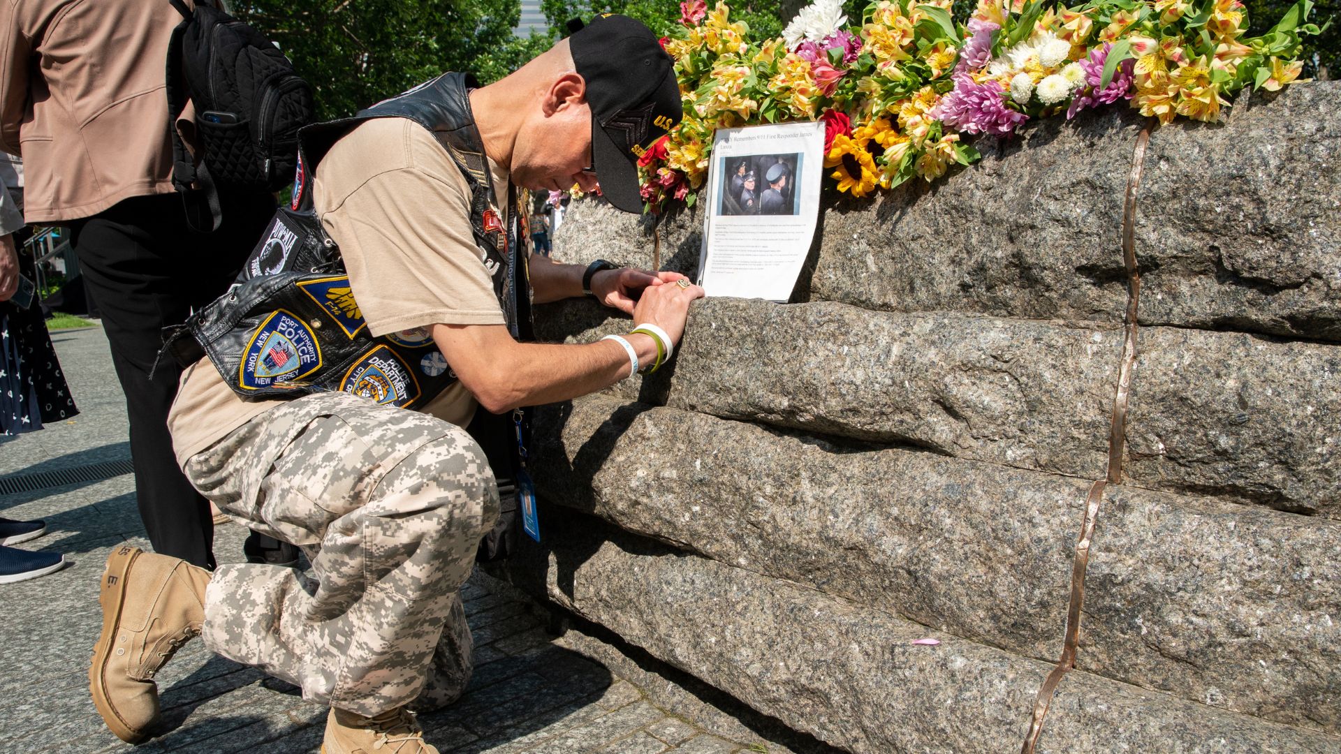A man wearing camouflage kneels at the Glade near tribute flowers and a photograph 