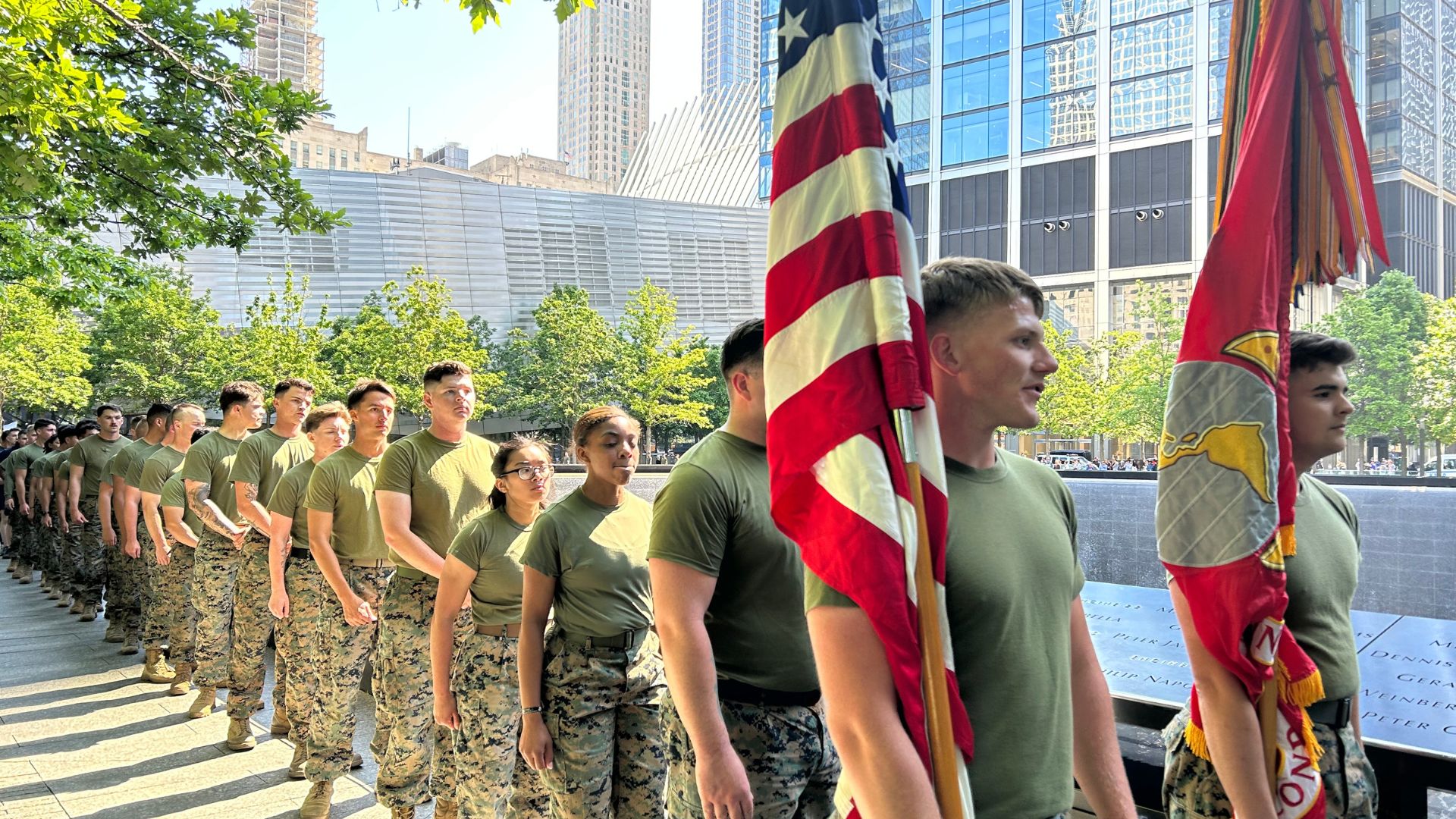 Soldiers in camouflage line up along the Memorial pools of the Memorial, with two at the front carrying flags.