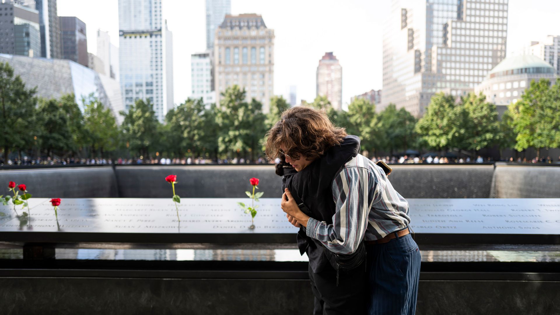Two people hug in front of the 9/11 Memorial, where there are several roses placed at victims' names, with part of the Manhattan skyline in background