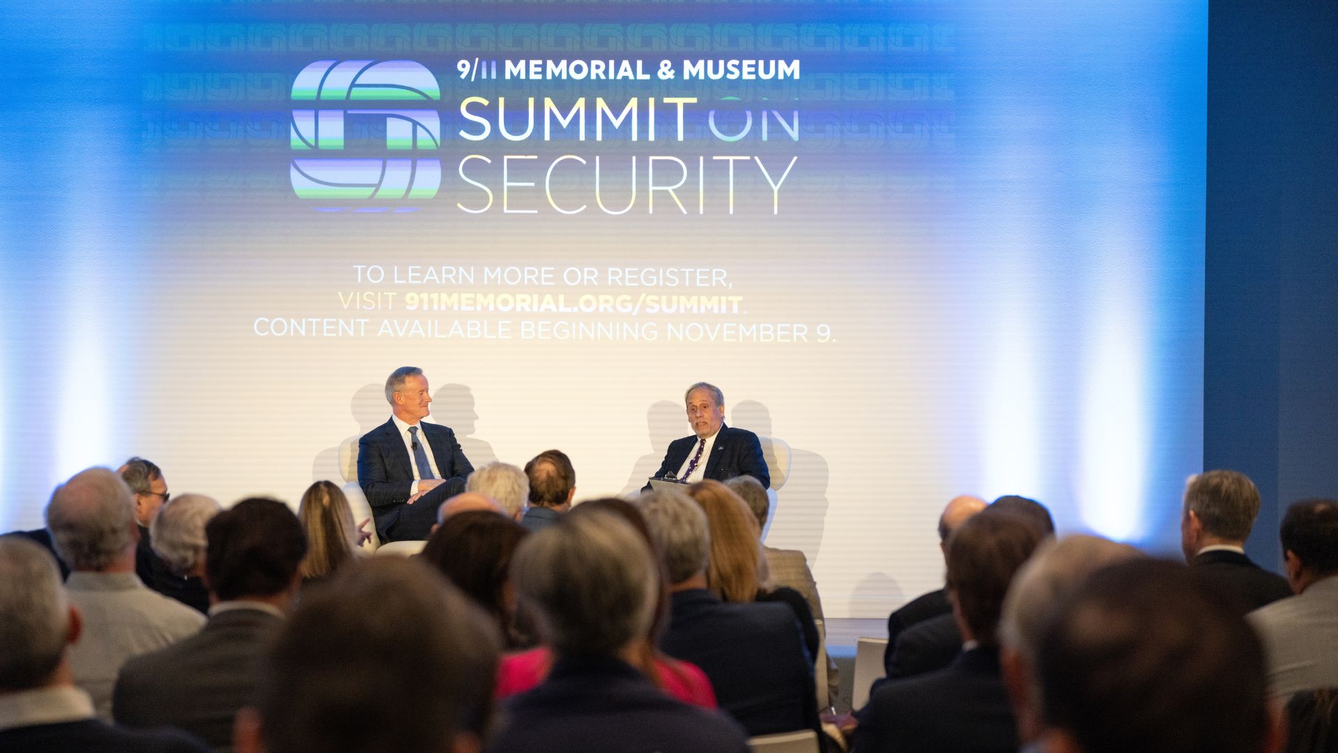 Back view of an audience listening to two speakers on stage, in front of a curtain with the 9/11 Memorial & Museum Summit on Security logo