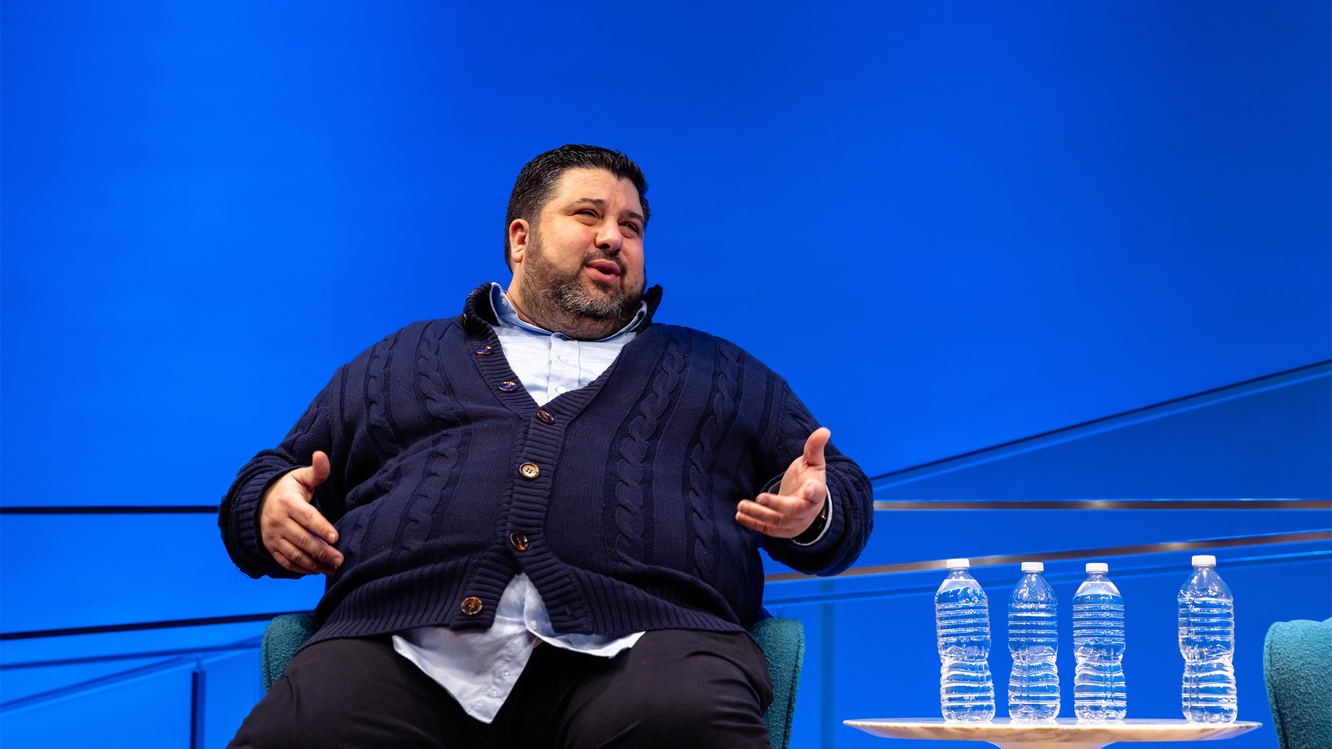 A bearded man wearing a cardigan sits in fron of a blue background.