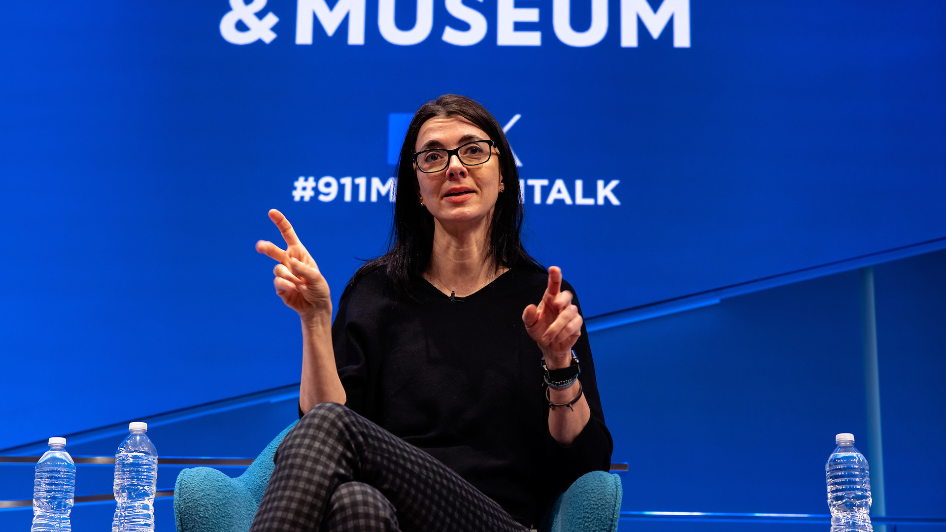 A woman with dark hair and glasses speaks on stage in front of a blue background displaying the 9/11 Memorial and Museum logo.