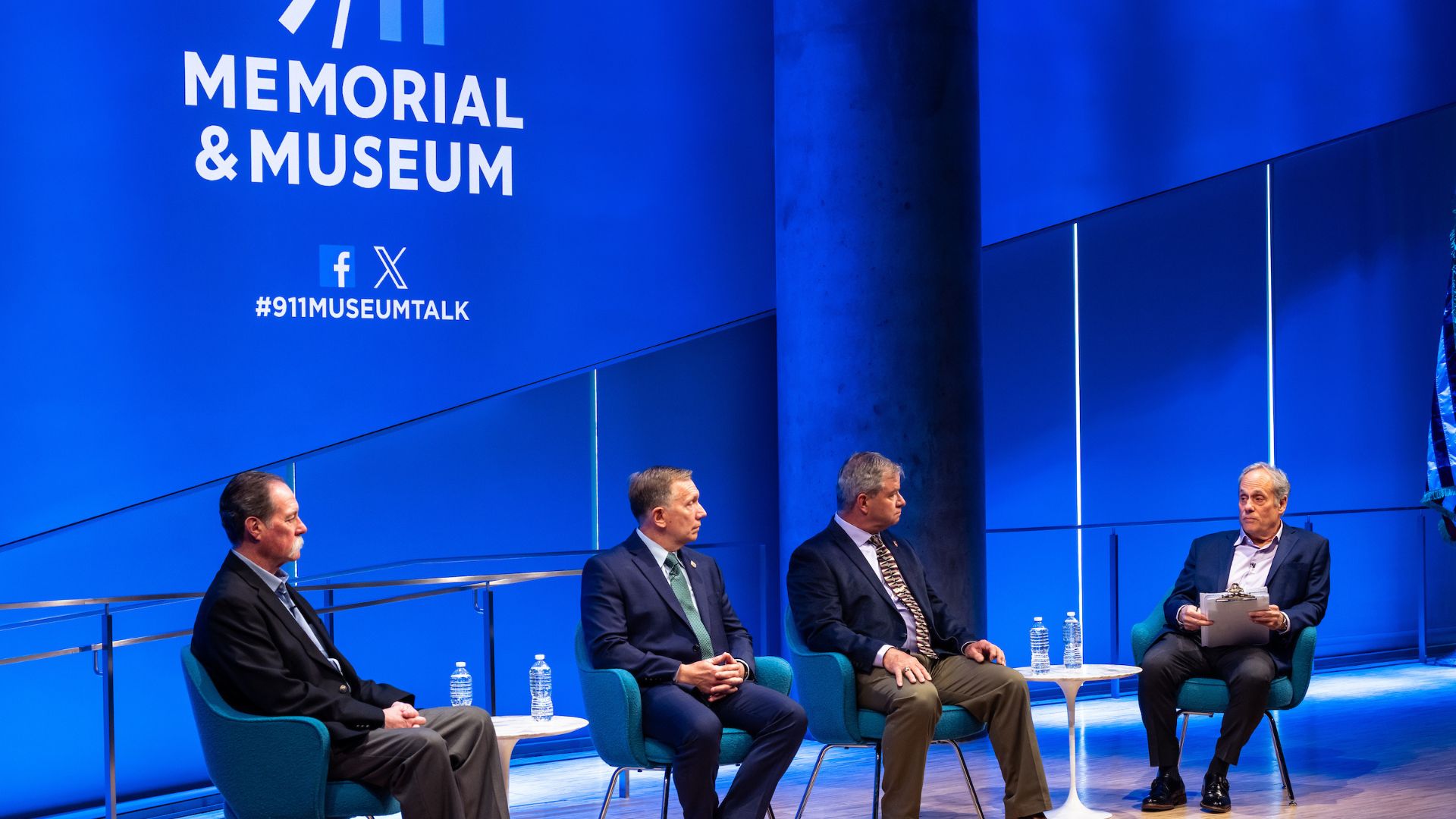 Panelists on stage against a bright blue curtain and the 9/11 Memorial & Museum logo.
