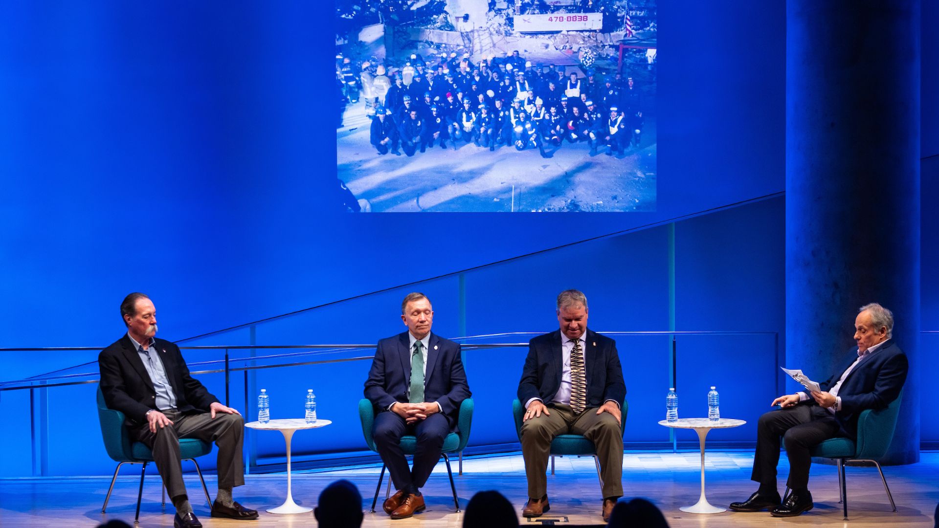 Panelists on stage against a bright blue curtain and the 9/11 Memorial & Museum logo.