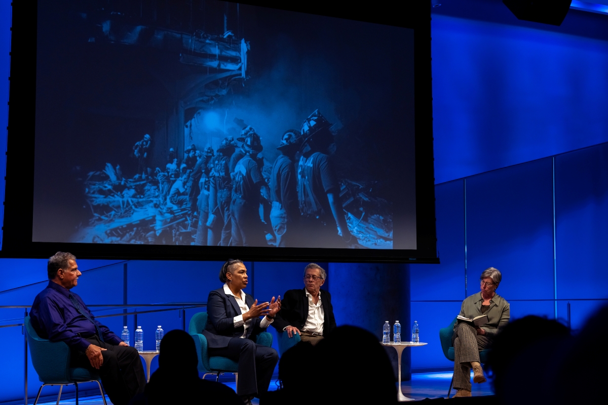 four people on stage sit in front of a large screen with a photo of rescue and recover efforts 