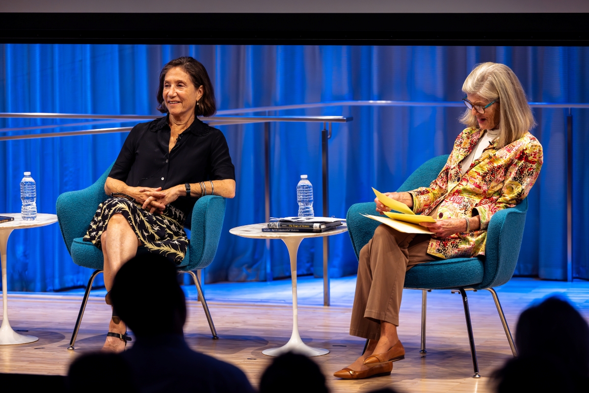 The host in a light jacket and a female speaker in black are pictured in front of blue curtains