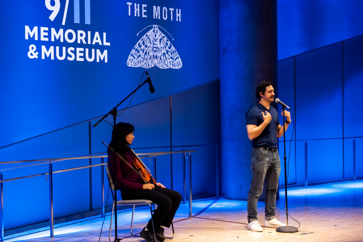 A Violinist sits on stage with her instrument and a man stands at the mic, gesturing, telling a story