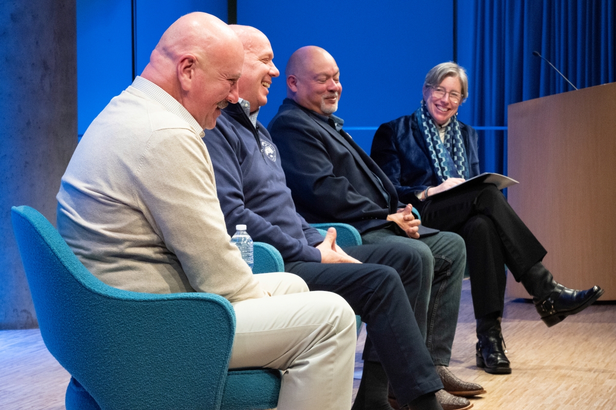 A group of three men and one woman speak on stage, smiling and laughing together.