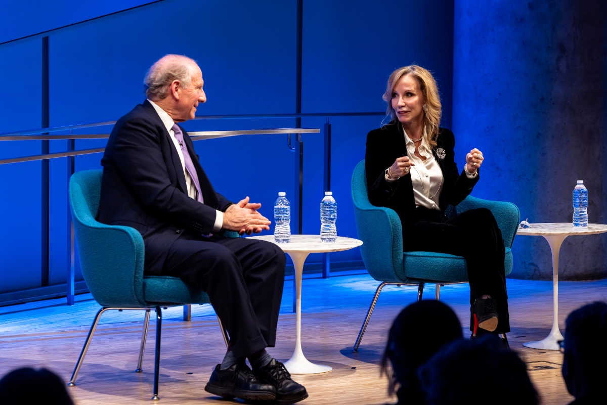 Two speakers sit on stage talking in front of a blue curtain