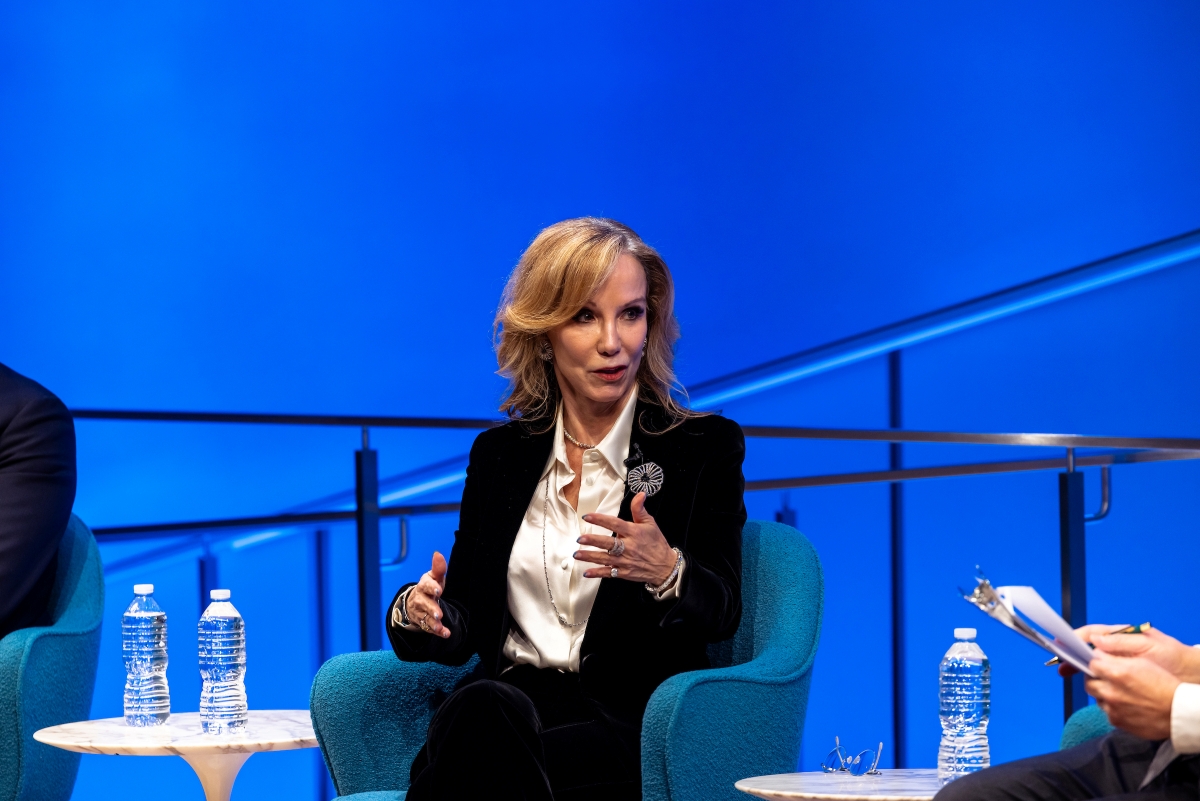 A woman with a dark jacket and white shirt gestures and speaks