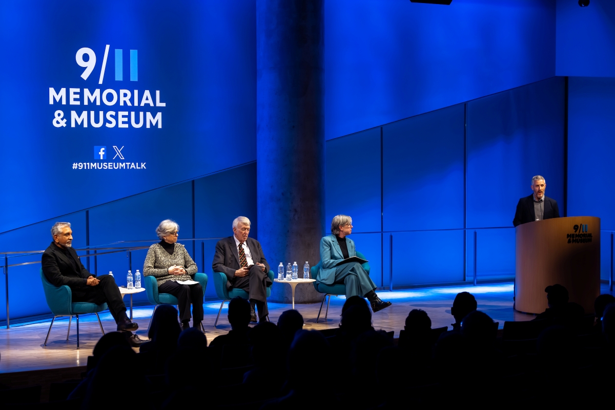 Three person panel sits on the stage with Beth Hillman, being introduced by a male speaker at the podium