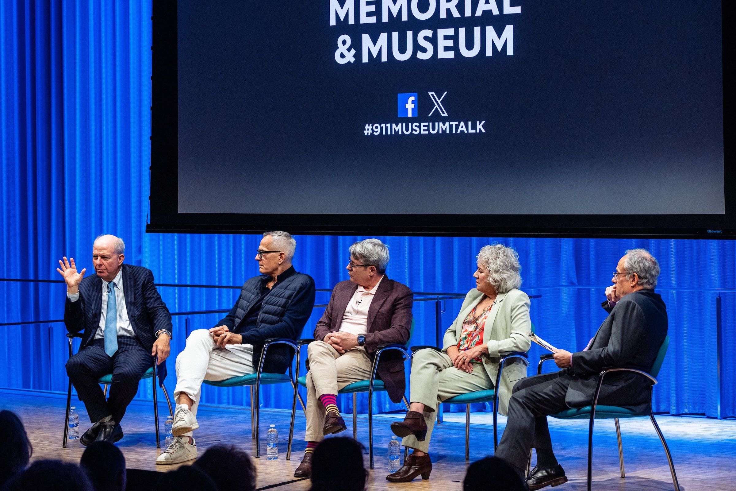 a man with a "five" gesture sits on stage speaking with four other people