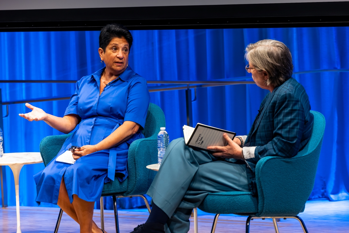 Two women are seated on stage having a conversation. A woman in a blue dress gestures with her right hand palm up and speaks to another woman, seen from a side profile with her head turned. The second woman listens, holding a notebook, wearing a green pantsuit in front of a blue curtain.