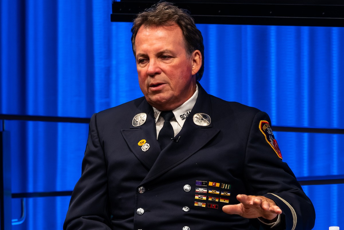 A man in an FDNY uniform gestures palm down and speaks from a seated position in front of a blue curtain.