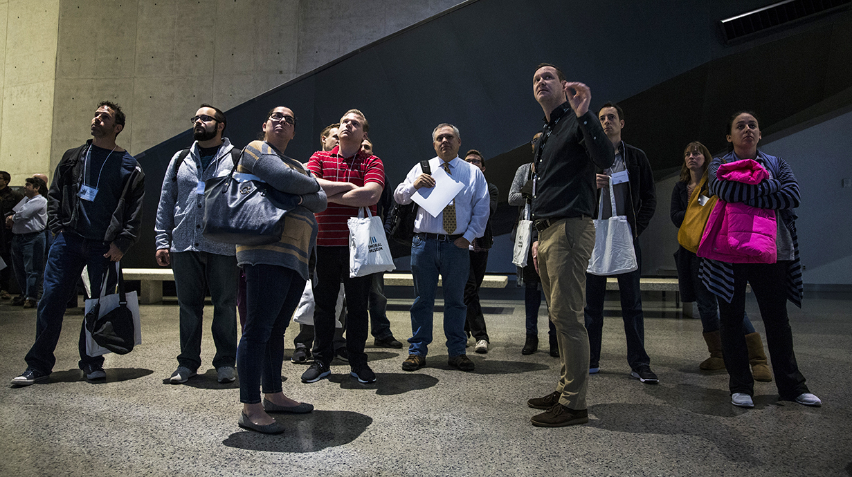 An instructor leads a teacher workshop at the Museum. He and a group of men and women are standing and facing forward as they observe an artifact. 