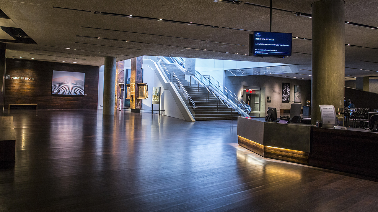 Sunlight pours down a large staircase into an empty Concourse Lobby. The light reflects off hardwood floors. An information desk is to the right and an illuminated image of the Twin Towers is off to the left. 