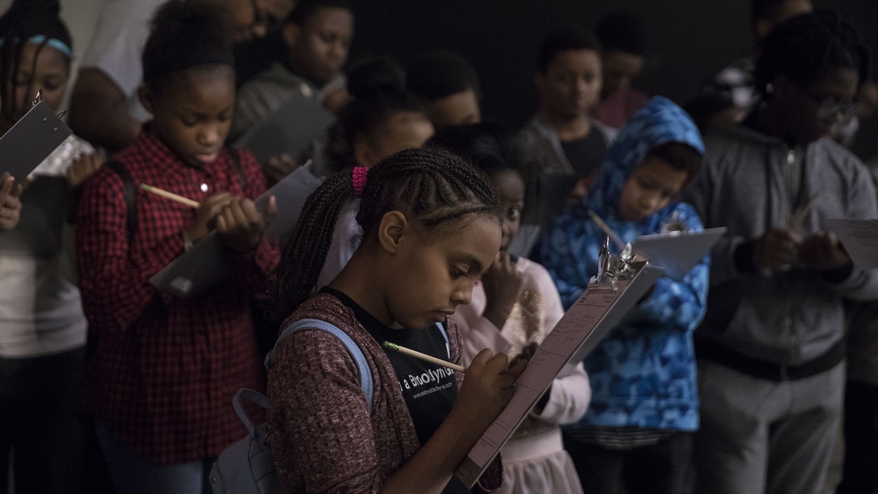 A student with braided hair writes on a clipboard as a group of her classmates do the same behind her. 