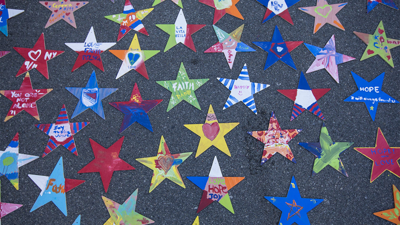 Dozens of hand-painted, colorful paper stars are spread out on a gray surface. Children have colored various 9/11-themed tributes on each star. The more than thirty stars contain messages that read, “hope,” “love,” and “I heart New York,” among others. 