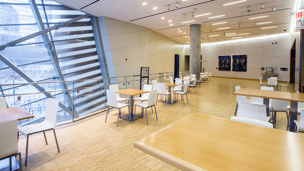 Small tables and chairs fill an empty atrium terrace at the museum Pavilion. The floors are hardwood and several pieces of art decorate the walls. 