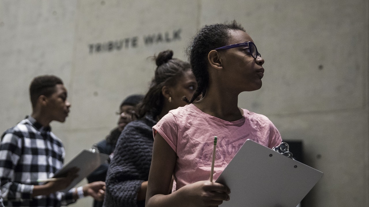 Several students with clipboards walk through the Tribute Walk, a Museum space where art related to 9/11 is displayed. 