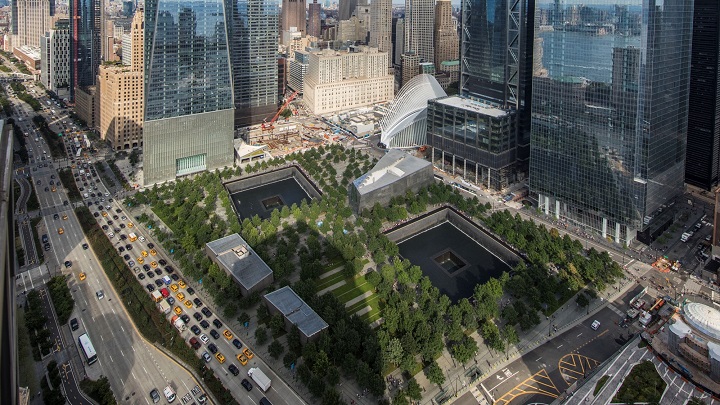Memorial Plaza is seen from above. The aerial view shows the Plaza’s many trees, as well as the reflecting pools and Museum Pavilion. Rows of traffic pass on West Street to the left of the Plaza. The buildings of lower Manhattan are in the background. 