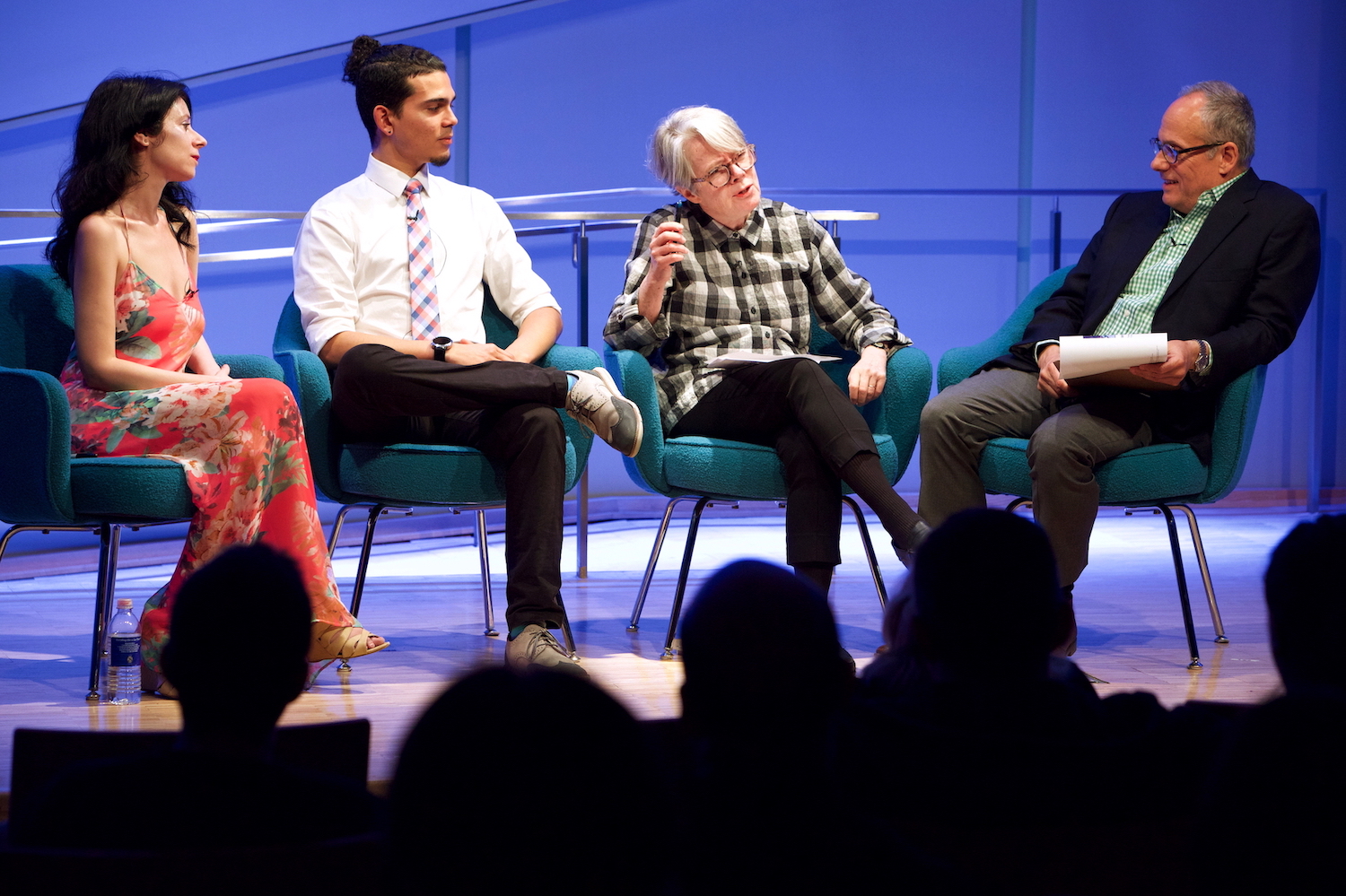 Four public program participants sit on a blue auditorium stage. A woman wearing glasses and a checked shirt is gesturing while speaking. The audience appears in the foreground in silhouette. 