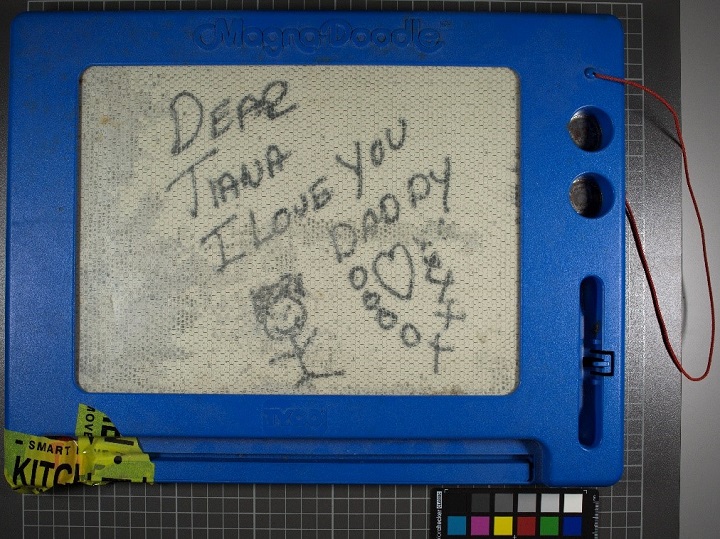 A close-up photograph of a message inscribed on a Magna Doodle with a blue plastic frame. On the Magna Doodle screen is a smiling stick figure and a message: "Dear Tiana / I Love You / Daddy." Below the message is a drawing of a heart surrounded by x's and o's. 