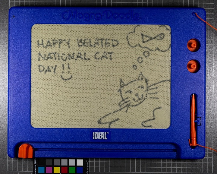 A message and a drawing inscribed on a Magna Doodle that appears slightly newer than the Magna Doodle in the previous image. On this screen is a drawing of a smiling cat reclining, a thought bubble featuring a fish above its head, and a message: "Happy Belated National Cat Day!!" with a smiley face. 