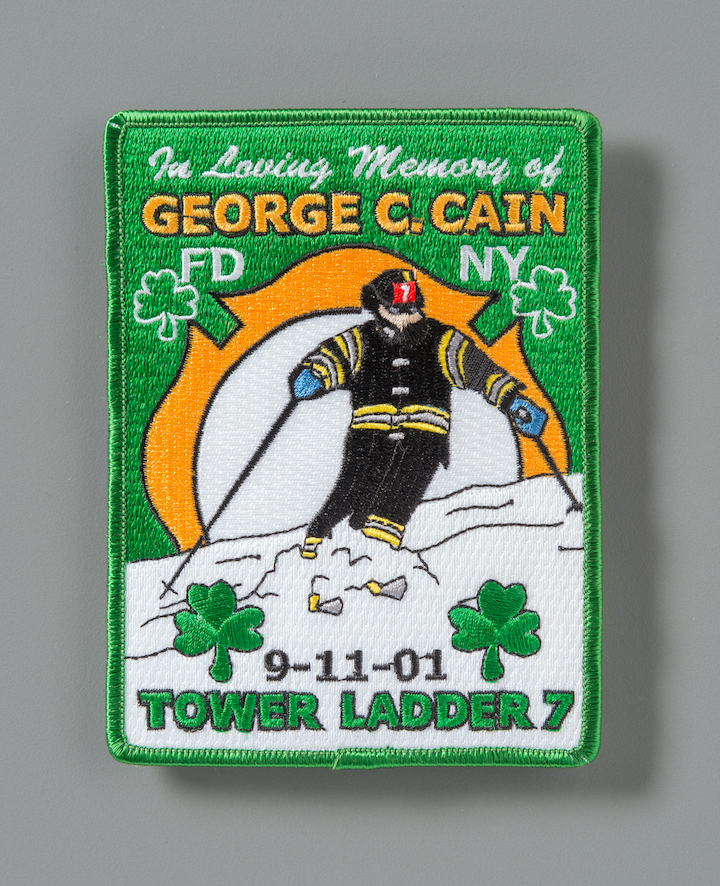 An embroidered memorial patch created in memory of FDNY firefighter George Cain depicts an illustration of a firefighter in bunker gear skiing in the snow. Two green shamrocks border the words Tower Ladder Seven. 